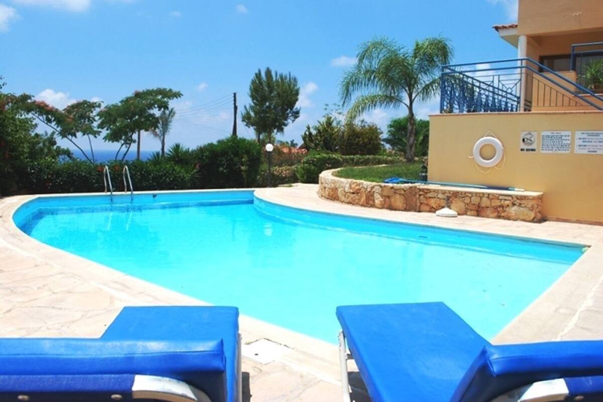 A swimming pool with blue lounge chairs, surrounded by stone walls and greenery, with a clear view of the ocean in the background.