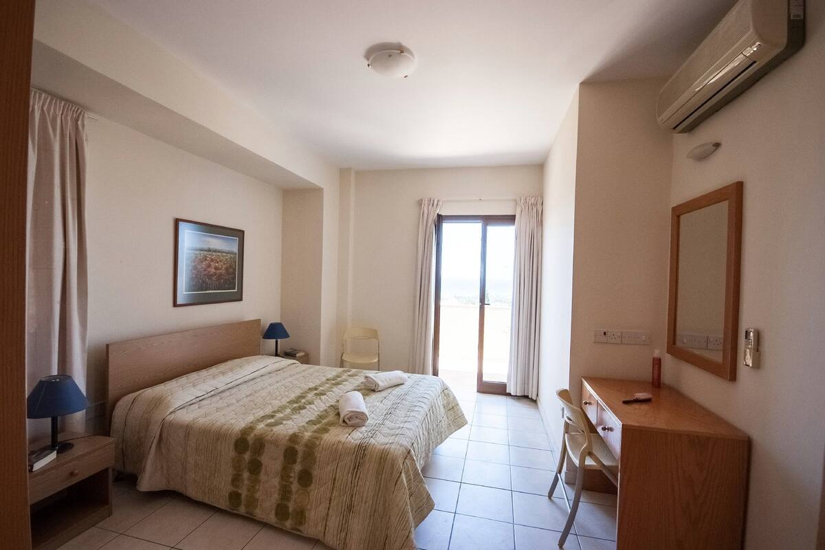 A hotel room with a bed, a desk, a chair, a window with curtains, and a picture on the wall.