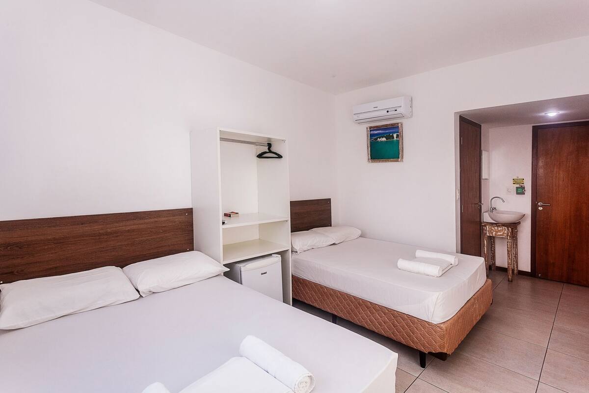 A hotel room with two single beds, a wardrobe, a small desk, a sink, and an air conditioning unit.