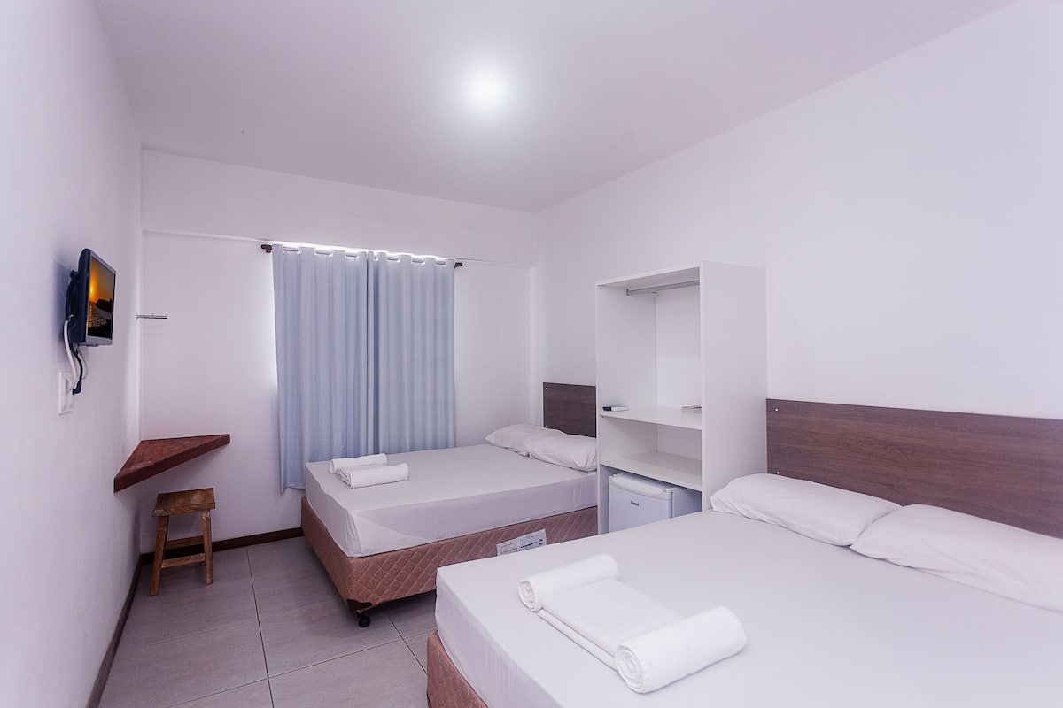 A hotel room with two beds, a small wooden stool, a TV mounted on the wall, and a window with curtains.