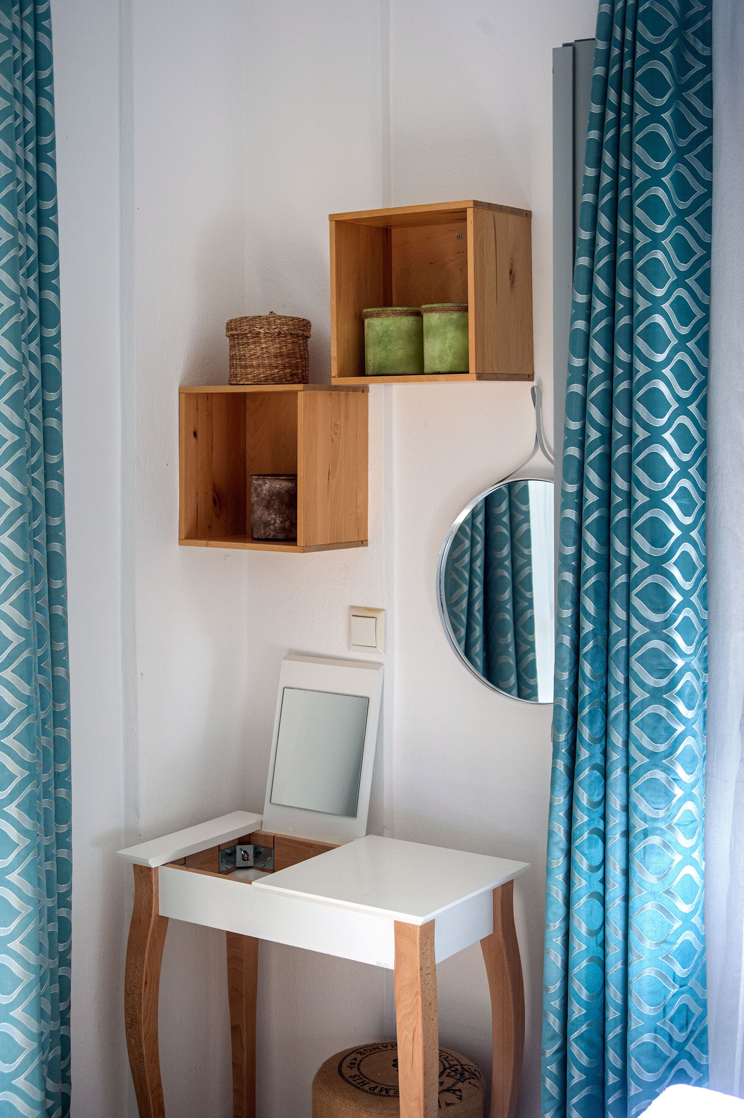 A small, white desk with a mirror above it, wooden shelves above the desk, and a patterned curtain to the right.