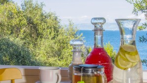 Une table avec une tasse, un bocal en verre, une bouteille rouge et un pichet dans lequel on a mis une tranche de citron, le tout placé devant un paysage pittoresque avec la mer et la verdure en toile de fond.