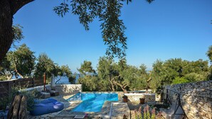Une piscine en pierre avec un élément décoratif aquatique bleu, entourée de plantes en pot et d’une chaise suspendue.