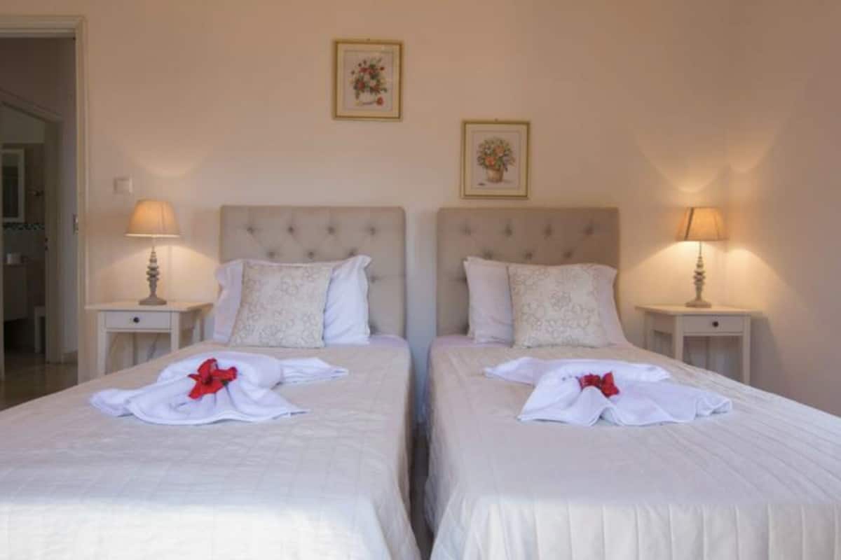 A hotel room with two beds, each with a white bedspread and a red bow. There are two bedside tables with lamps, and the walls are adorned with framed floral artwork.