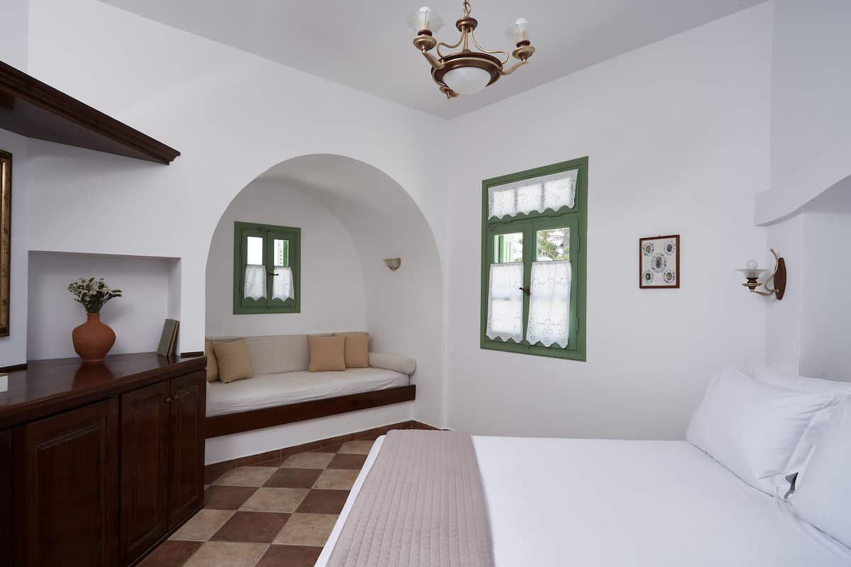 A bedroom with a bed, a sofa, a window with green shutters, a chandelier, and a wooden cabinet.