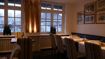 A dining area with a table set for two, chairs, a radiator, and a window with a view of snow-covered trees.