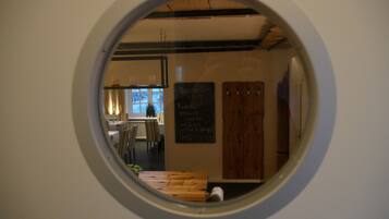 Une vue à travers une fenêtre circulaire donnant sur une pièce où se trouvent une table en bois, des chaises, un tableau noir servant de menu et une fenêtre avec des rideaux.