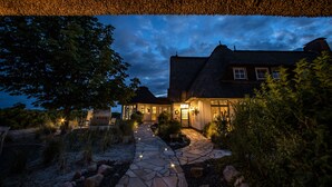 A stone pathway leading to a house with a thatched roof, surrounded by trees and shrubs. - Lundenbergsand Hotel und Spa (Simonsberg)