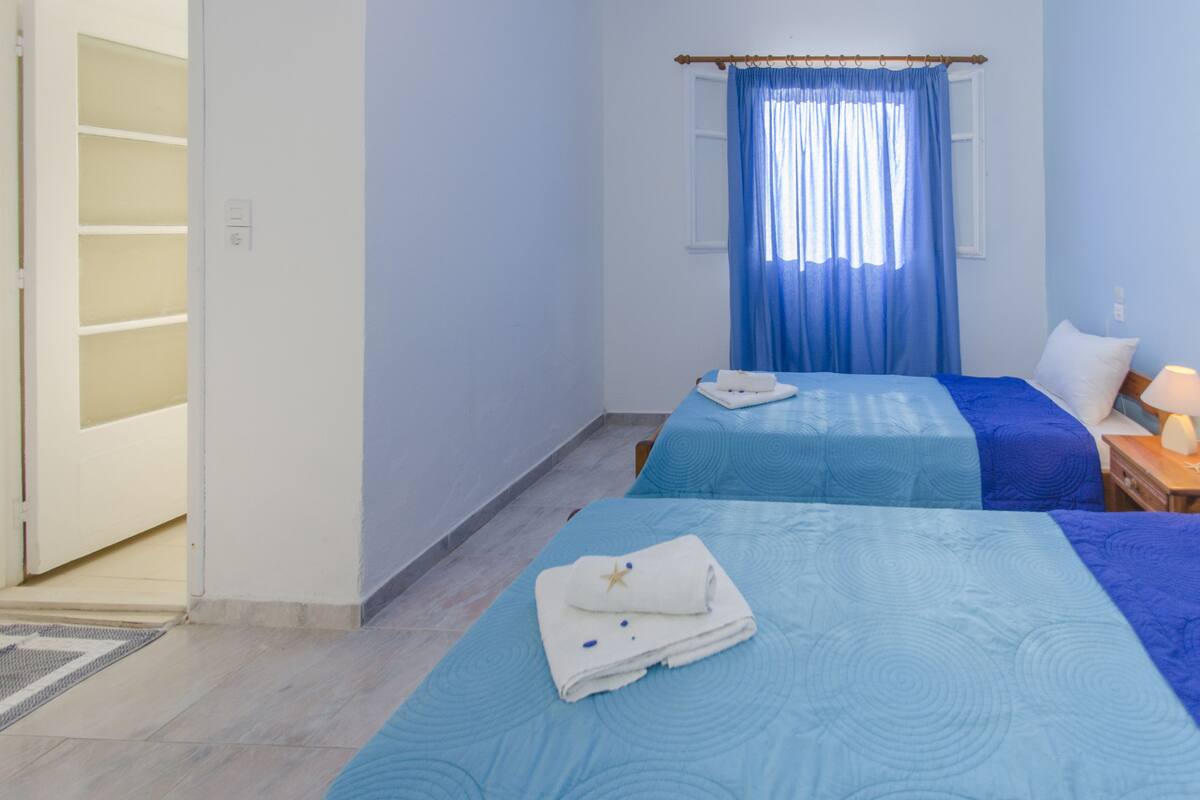 A hotel room with two beds, a window with blue curtains, a bedside table with a lamp, and a door leading to a storage area.