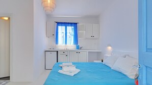 A bedroom with a bed, a small desk, a window with curtains, and a kitchenette. - Thalassopetra (Milos)