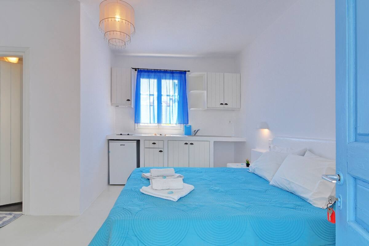 A bedroom with a bed, a small desk, a window with curtains, and a kitchenette.