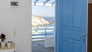 A room with a view of a beach and cliffs, a blue door, and a small table with a vase and cups. - Thalassopetra (Milos)