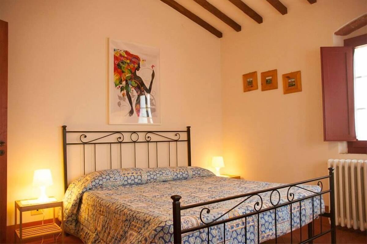 A bedroom with a bed, two bedside tables, a radiator, and a wall with a painting and three small framed pictures.