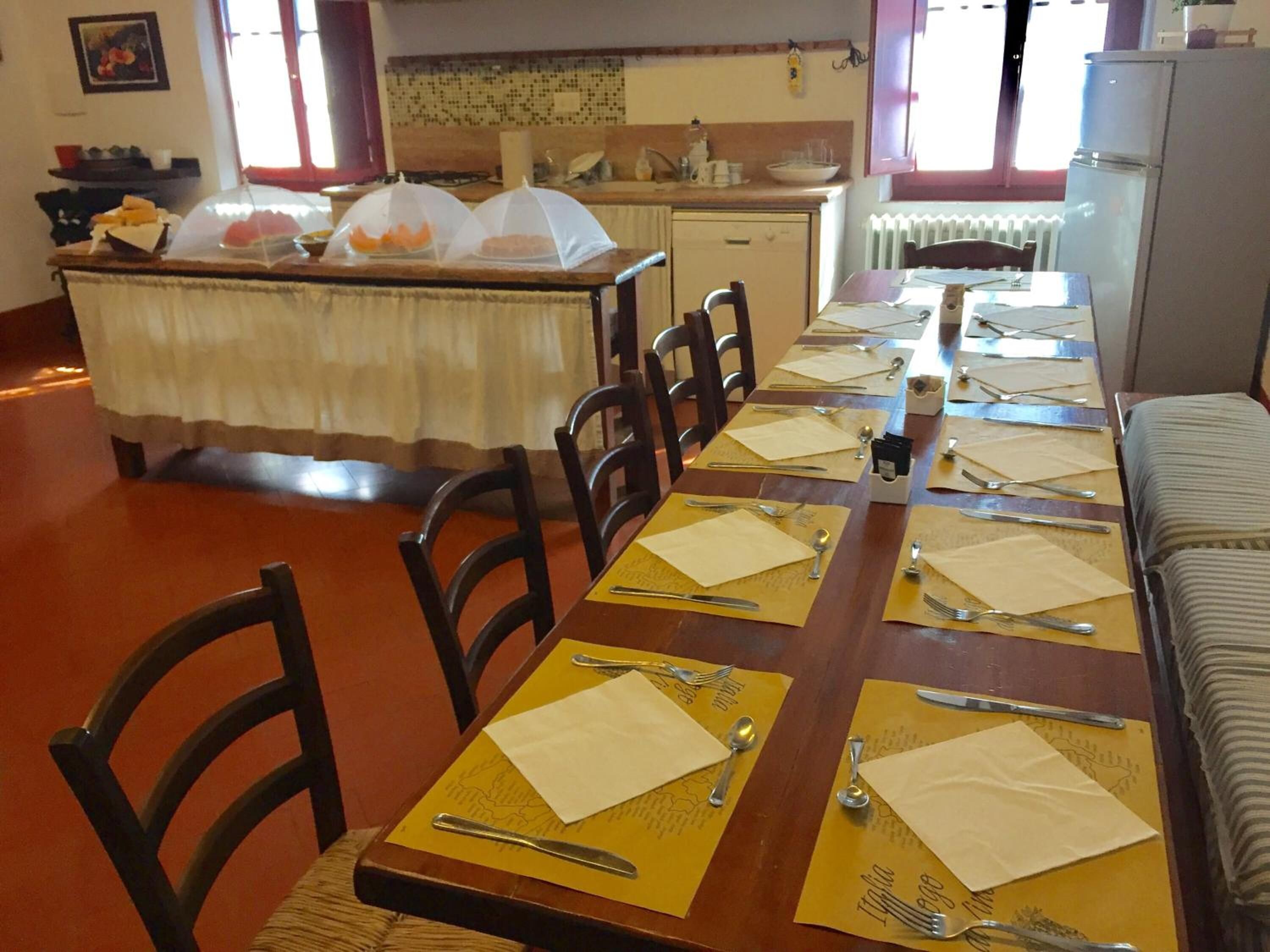 Un comedor con una mesa larga dispuesta para varias personas, sillas, una zona de cocina con refrigerador y una ventana con cortinas.