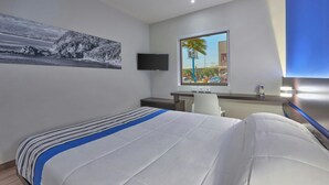 Quarto de hotel com cama, escrivaninha, cadeira, televisão e janela com vista para palmeiras e edifícios.