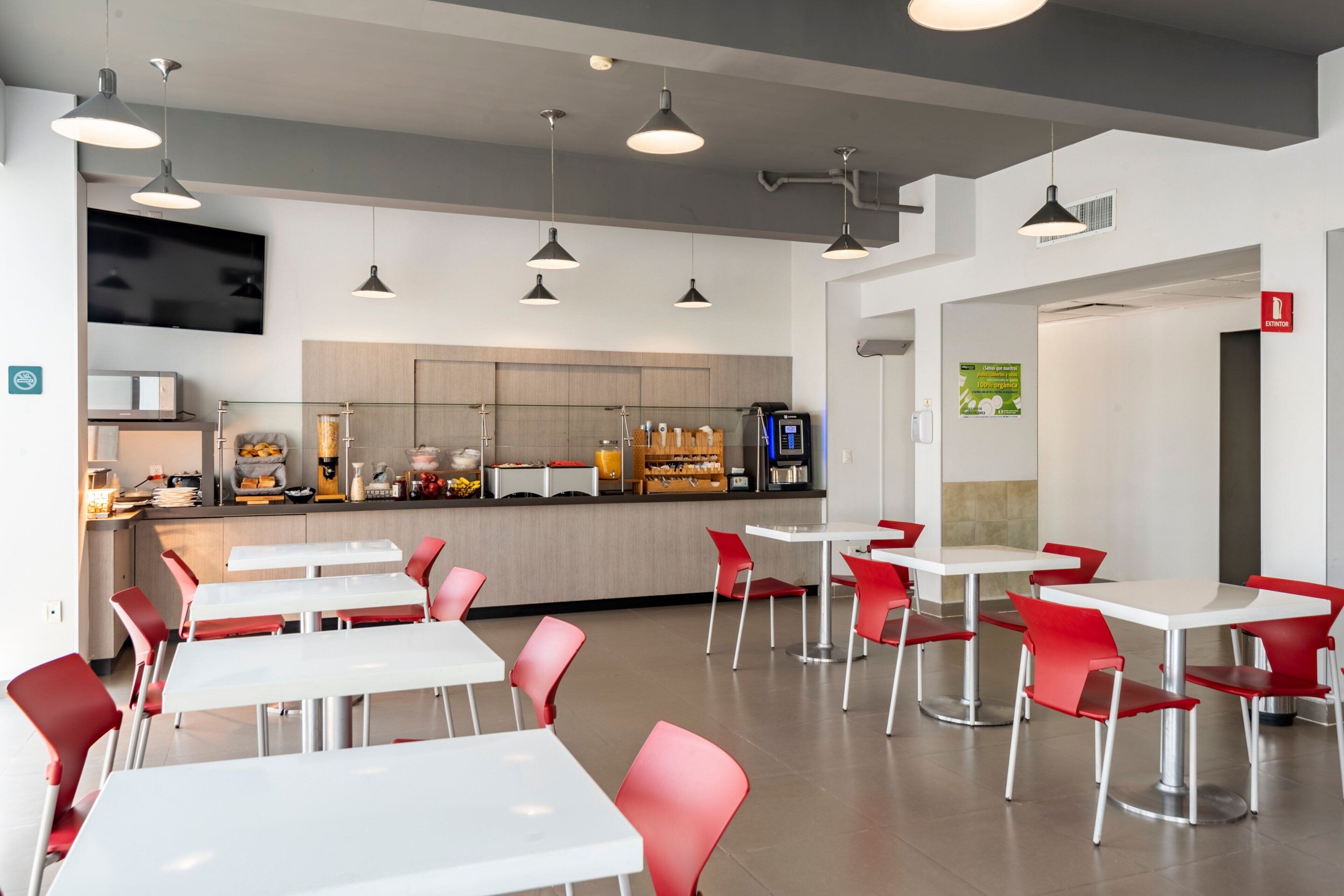 Una cafetería moderna con mesas blancas y sillas rojas, una zona de cocina con una máquina de café y un cartel que señala un contenedor de reciclaje.