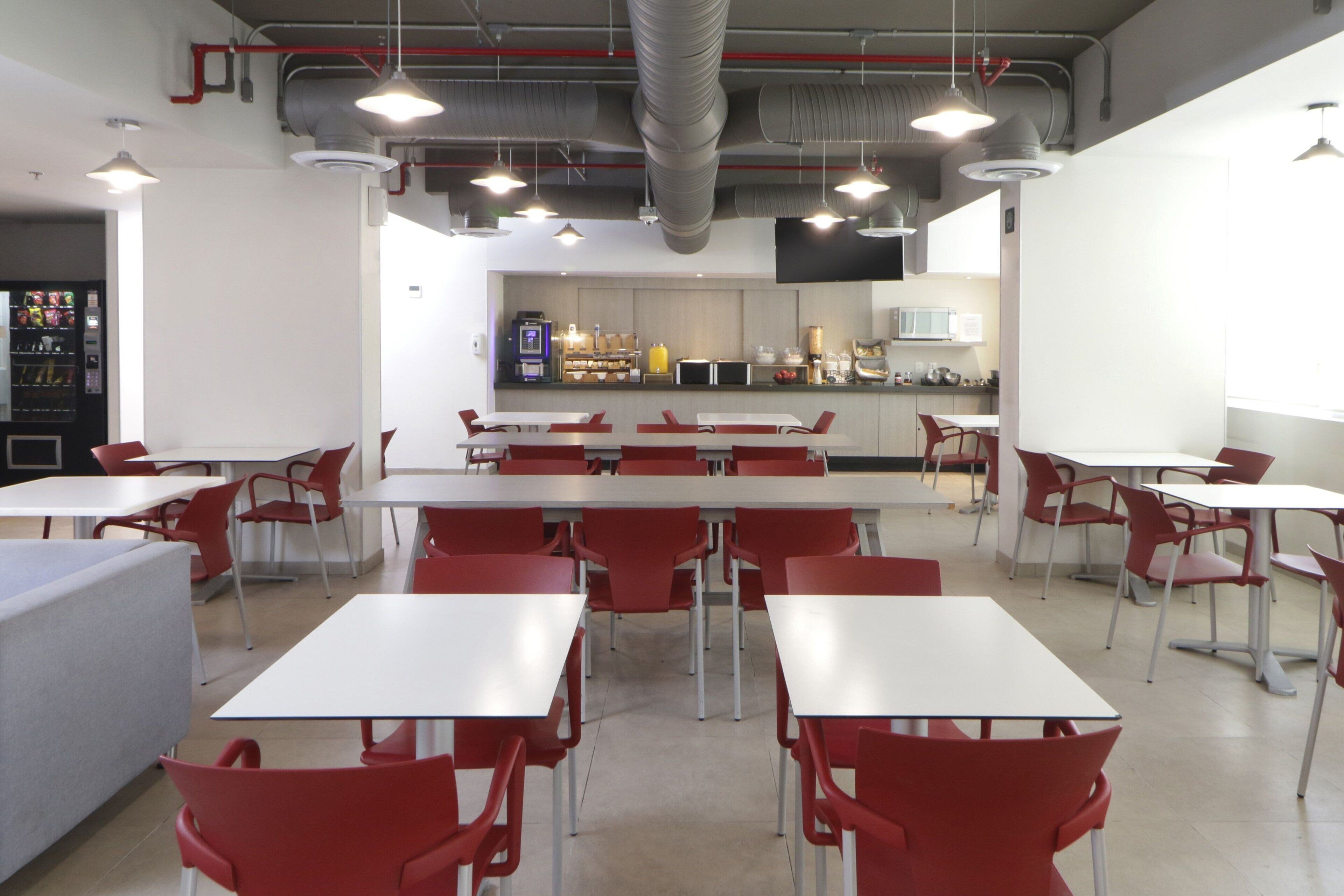 A modern cafeteria with white tables and red chairs, a visible kitchen area, and a vending machine in the background.
