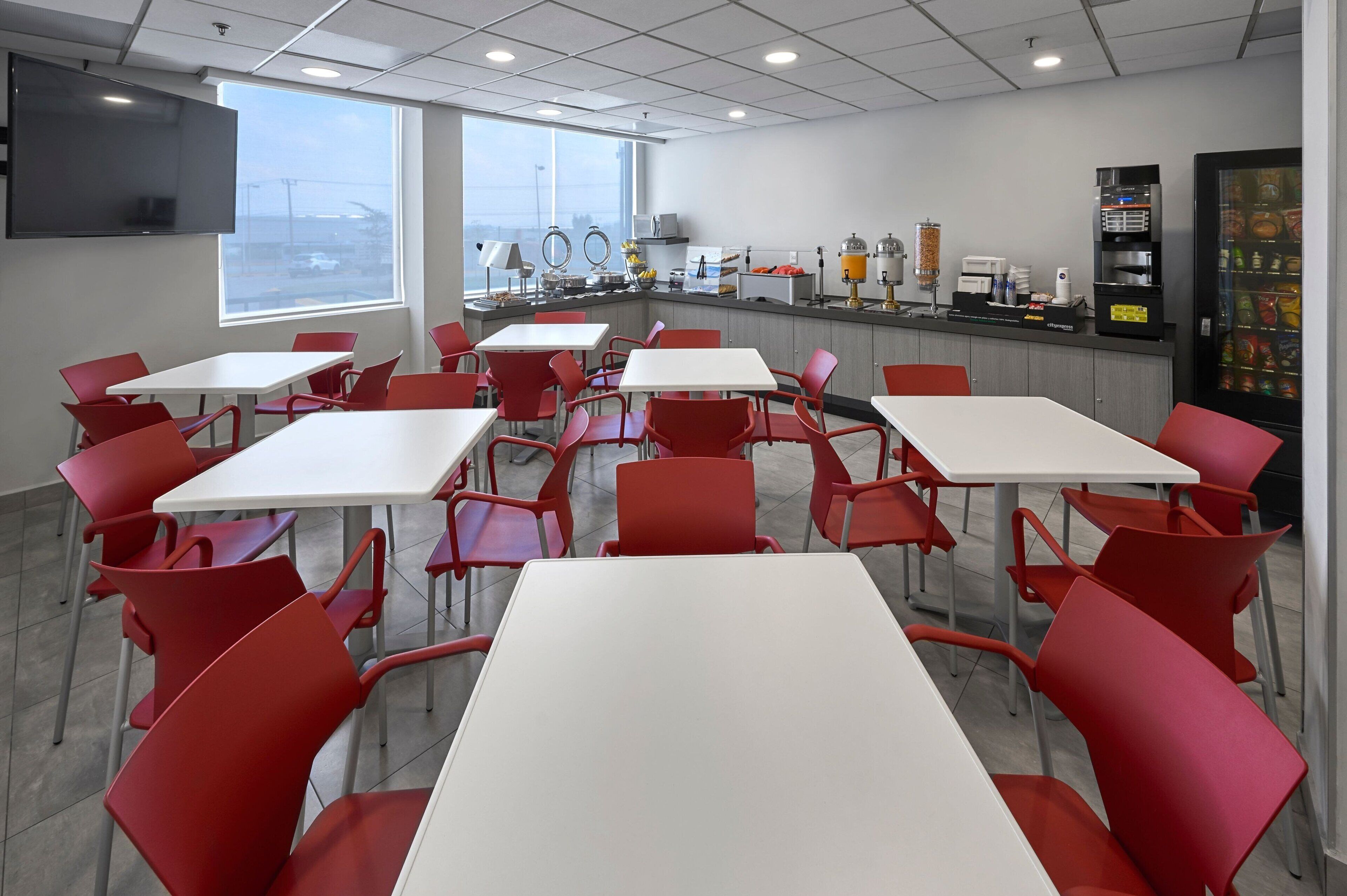 Uma zona de refeições moderna com cadeiras vermelhas e mesas brancas, bancada de cozinha com vários eletrodomésticos, e uma máquina de vendas automática.
