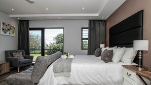 A modern bedroom with a large bed, a grey armchair, a nightstand, and a view of the outdoors. - Casa Ridge (Umhlanga)