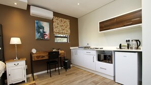 A compact kitchen with white cabinets, a microwave, a coffee maker, and a refrigerator. There's a wooden side table with a lamp and a chair. - Casa Ridge (Umhlanga)