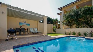 A swimming pool with a tiled deck, a barbecue grill, and a patio area with outdoor furniture. - Casa Ridge (Umhlanga)
