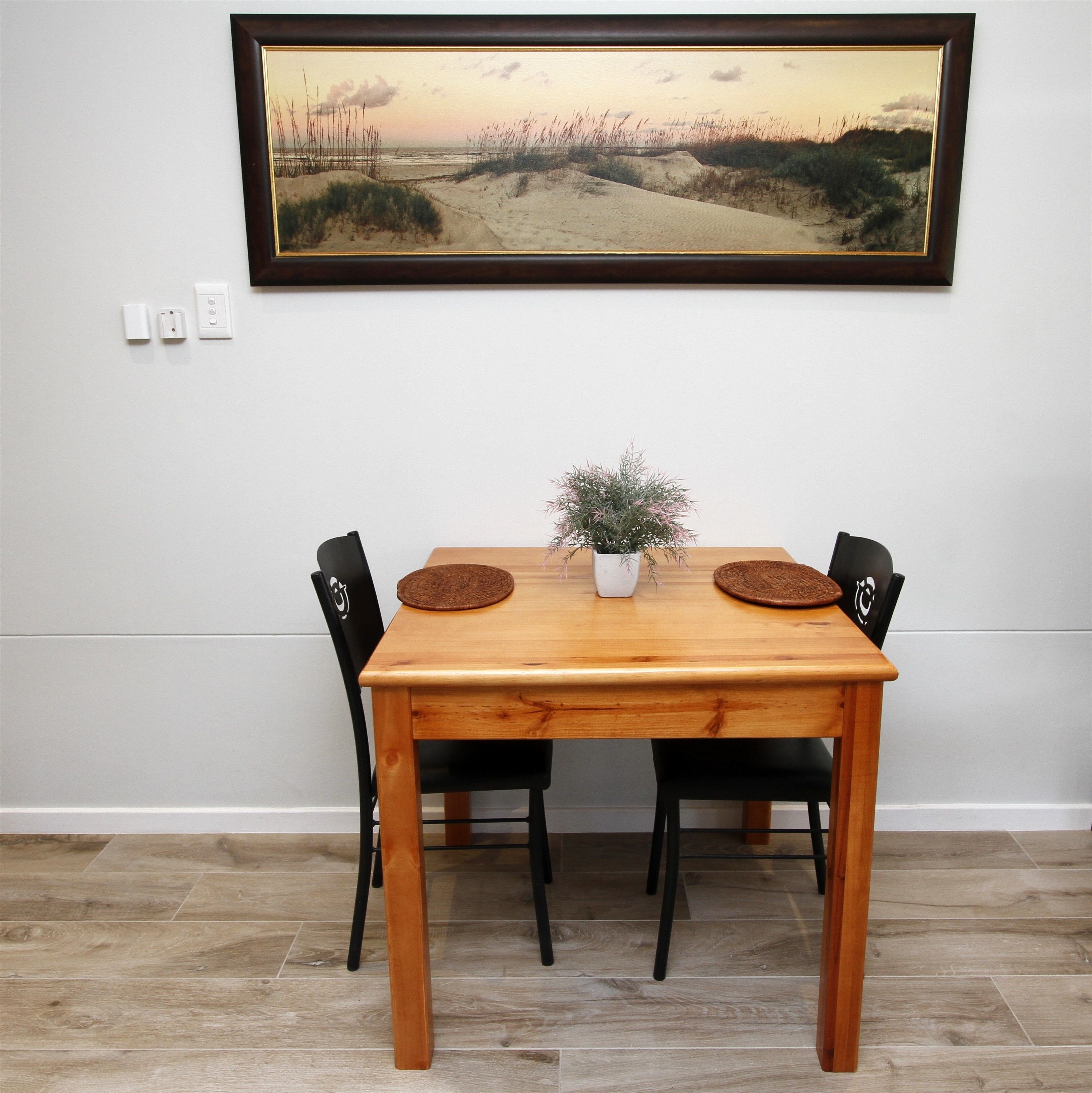 Uma mesa de jantar com quatro cadeiras, um vaso de planta e um quadro com paisagem na parede.