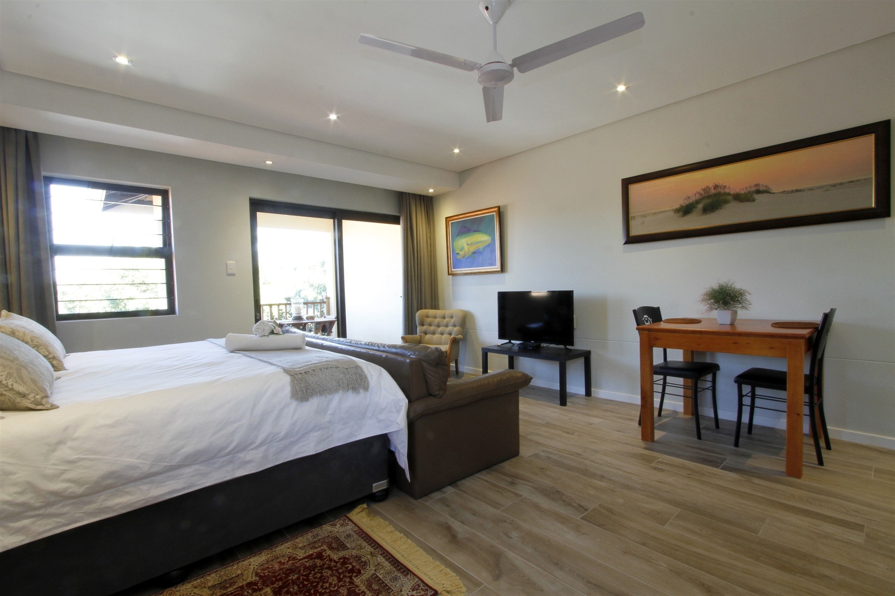 A bedroom with a bed, a sofa, a TV, a desk with chairs, and a painting on the wall.