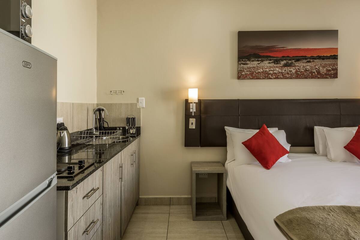 A hotel room with a bed, a nightstand, a small table, a kitchen area, and a painting on the wall.