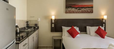 A hotel room with a bed, a nightstand, a small table, a kitchen area, and a painting on the wall.