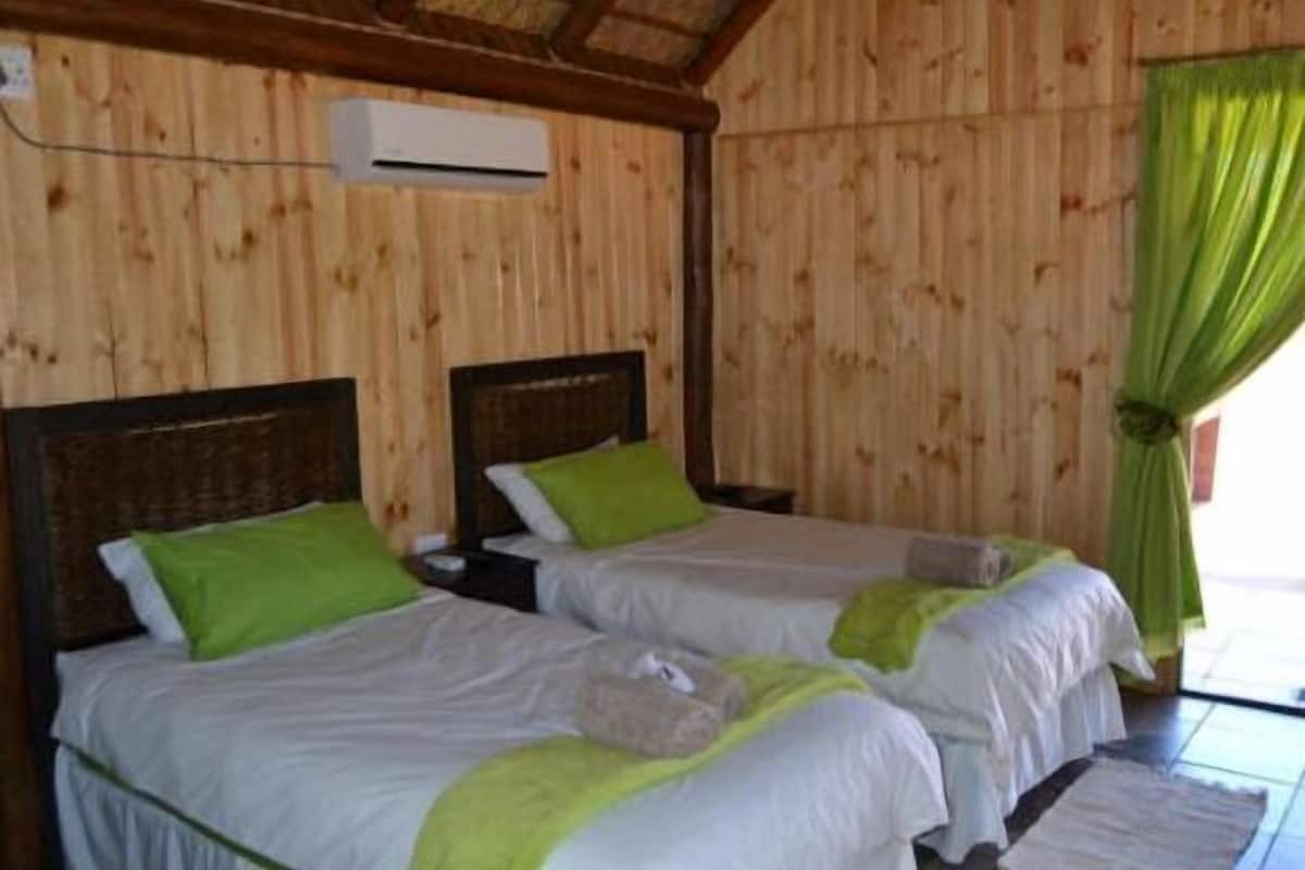 Een kamer met twee bedden, elk met een groen kussen en een witte sprei. Er is een airconditioningunit aan de muur boven de bedden bevestigd.