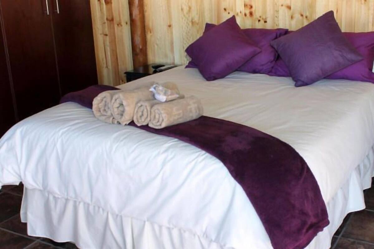 Een bed met een witte deken, paarse kussens en een opgerolde handdoek.
