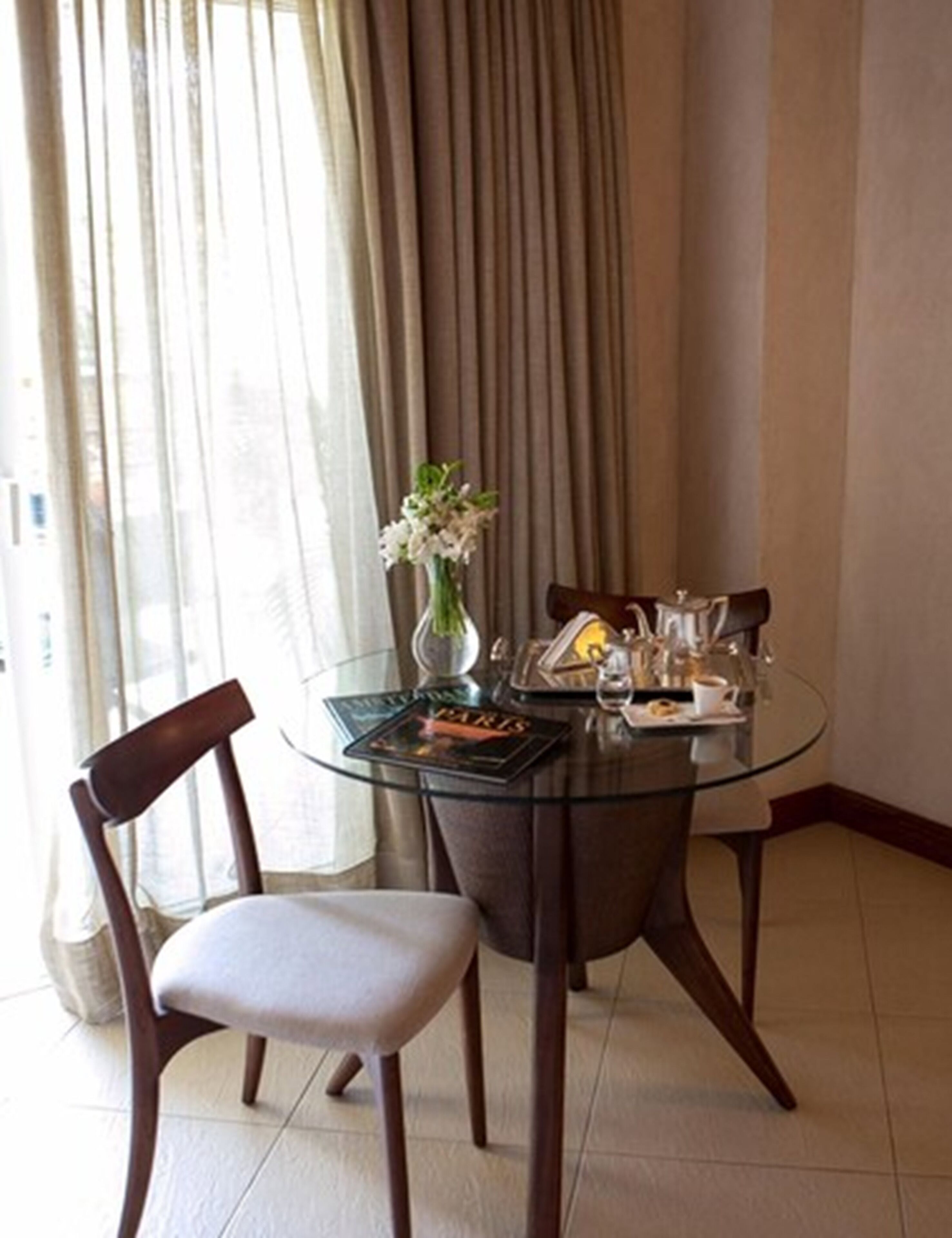 Un comedor con una mesa de vidrio, dos sillas, un florero con flores y una revista sobre la mesa.