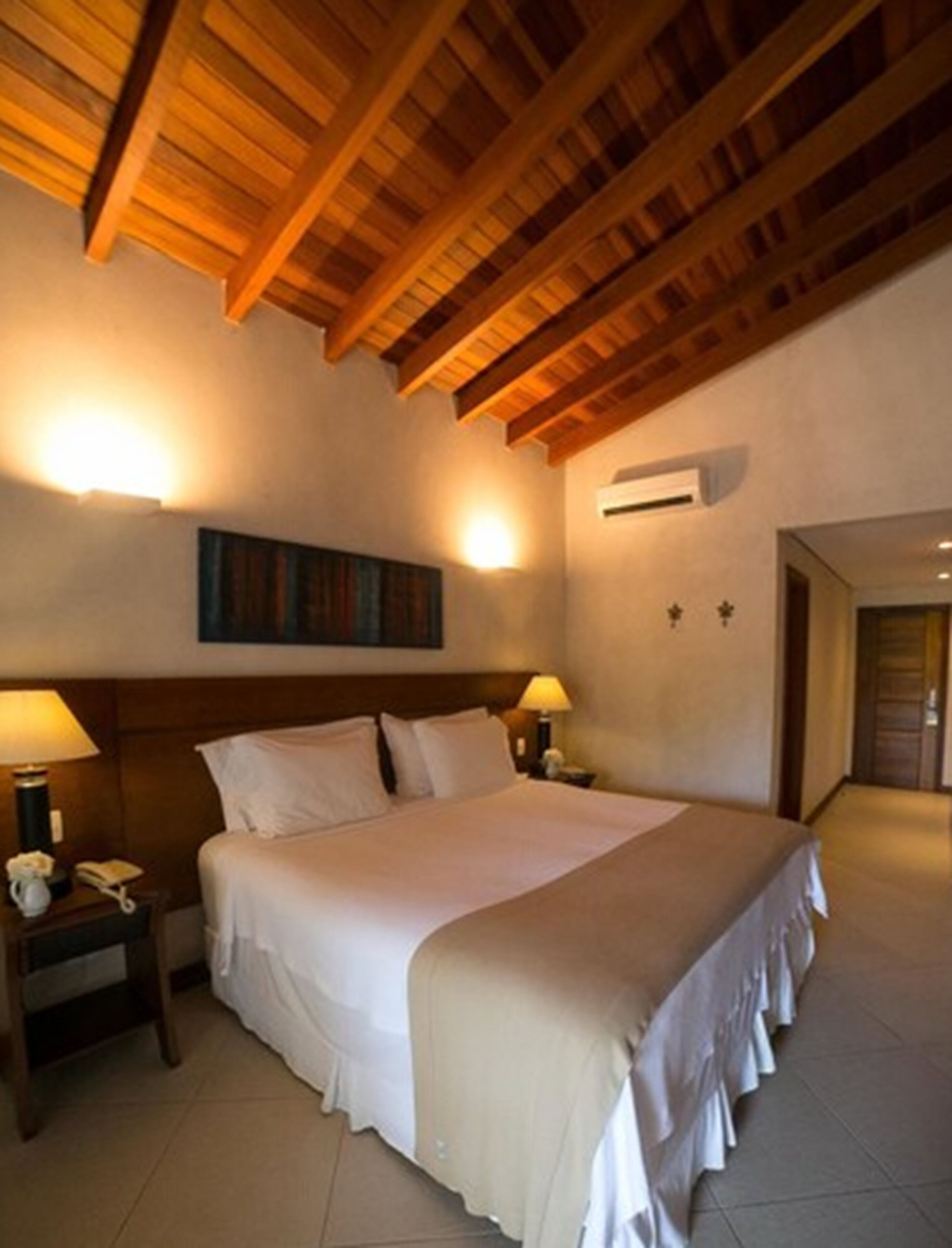 A hotel room with a wooden ceiling, a large bed with white bedding, two bedside tables with lamps, and an air conditioning unit on the wall.