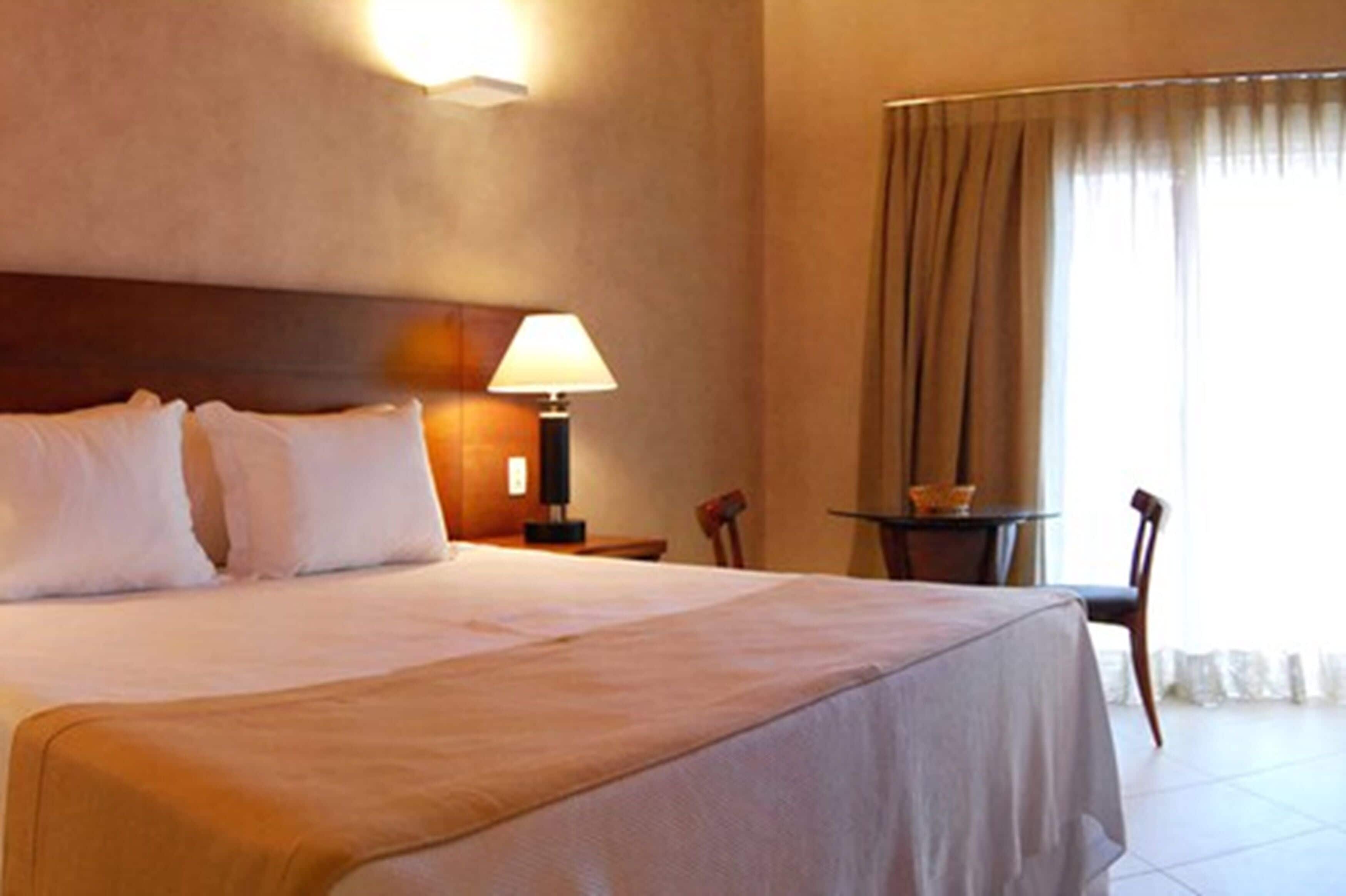 A hotel room with a large bed, a bedside table with a lamp, a small table with a basket, and a chair.