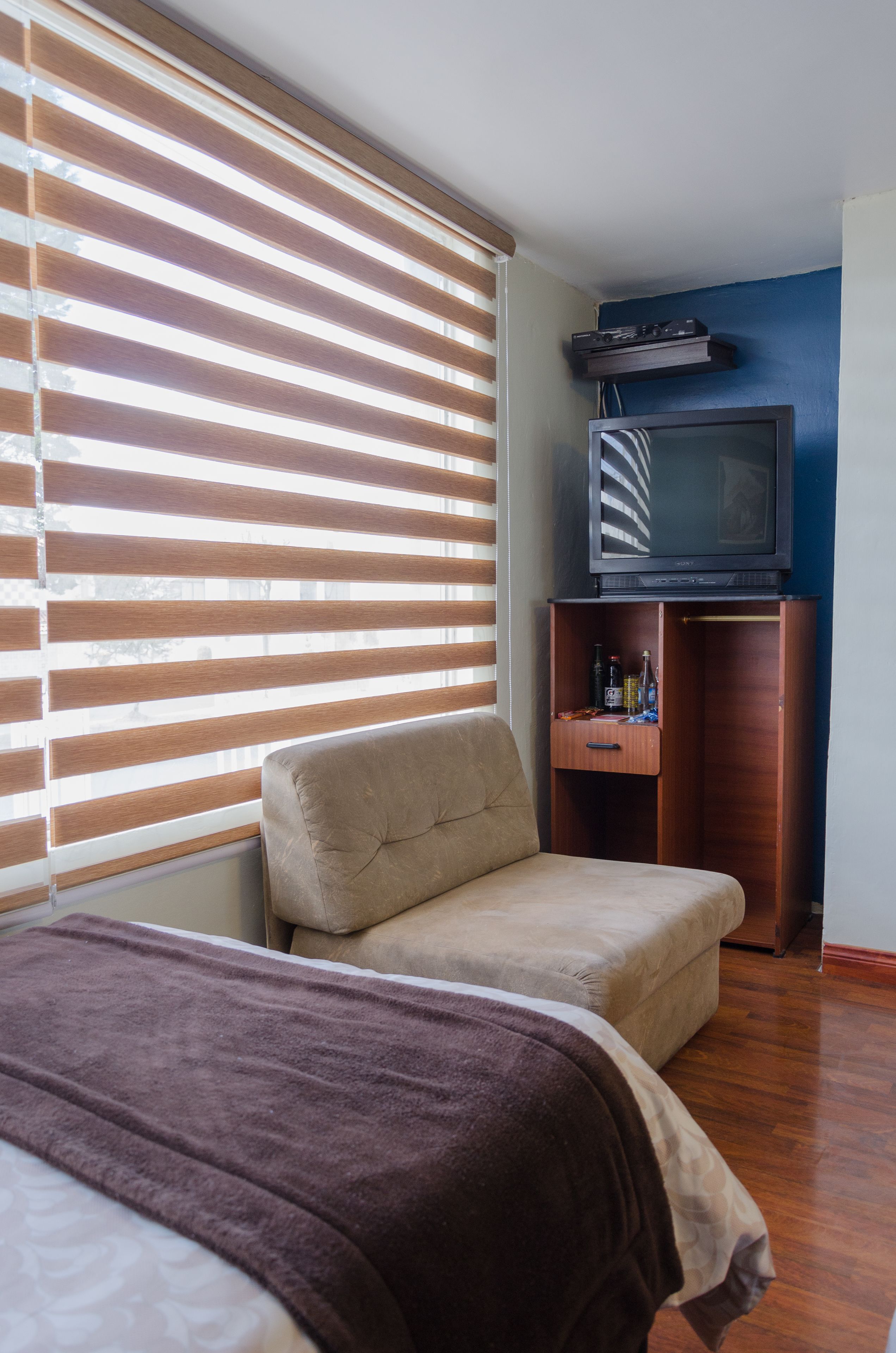 Una habitación con una cama, un sofá pequeño, un televisor sobre un mueble de madera y una ventana con persianas.