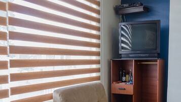 Una habitación con una cama, un sofá pequeño, un televisor sobre un mueble de madera y una ventana con persianas.
