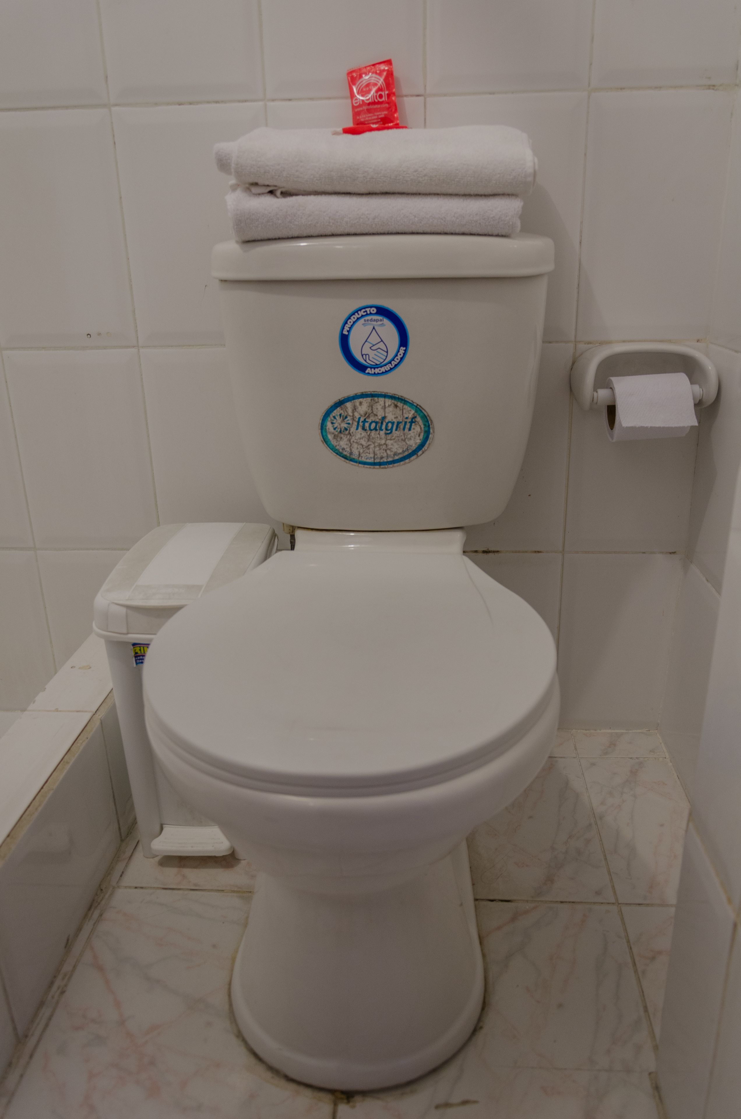 Un inodoro blanco con una calcomanía azul y blanca en el tanque, un soporte blanco para papel higiénico y un basurero blanco.