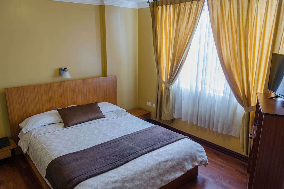 A hotel room with a wooden headboard, a bed with white and brown bedding, a bedside lamp, a TV on a wooden stand, and a window with curtains.