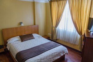 Una habitación de hotel con cabecera de madera, cama con ropa de cama blanca y marrón, lámpara de noche, televisor sobre un soporte de madera y una ventana con cortinas.