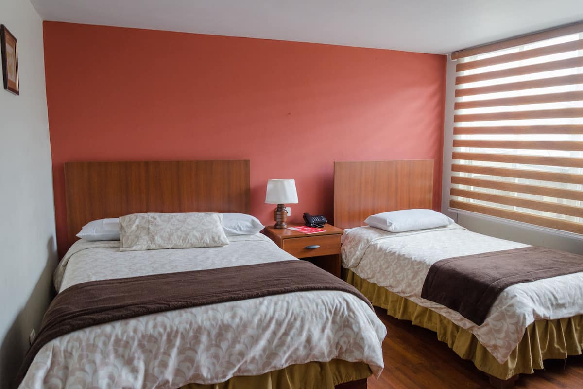 A hotel room with two beds, a nightstand with a lamp, and a window with blinds.