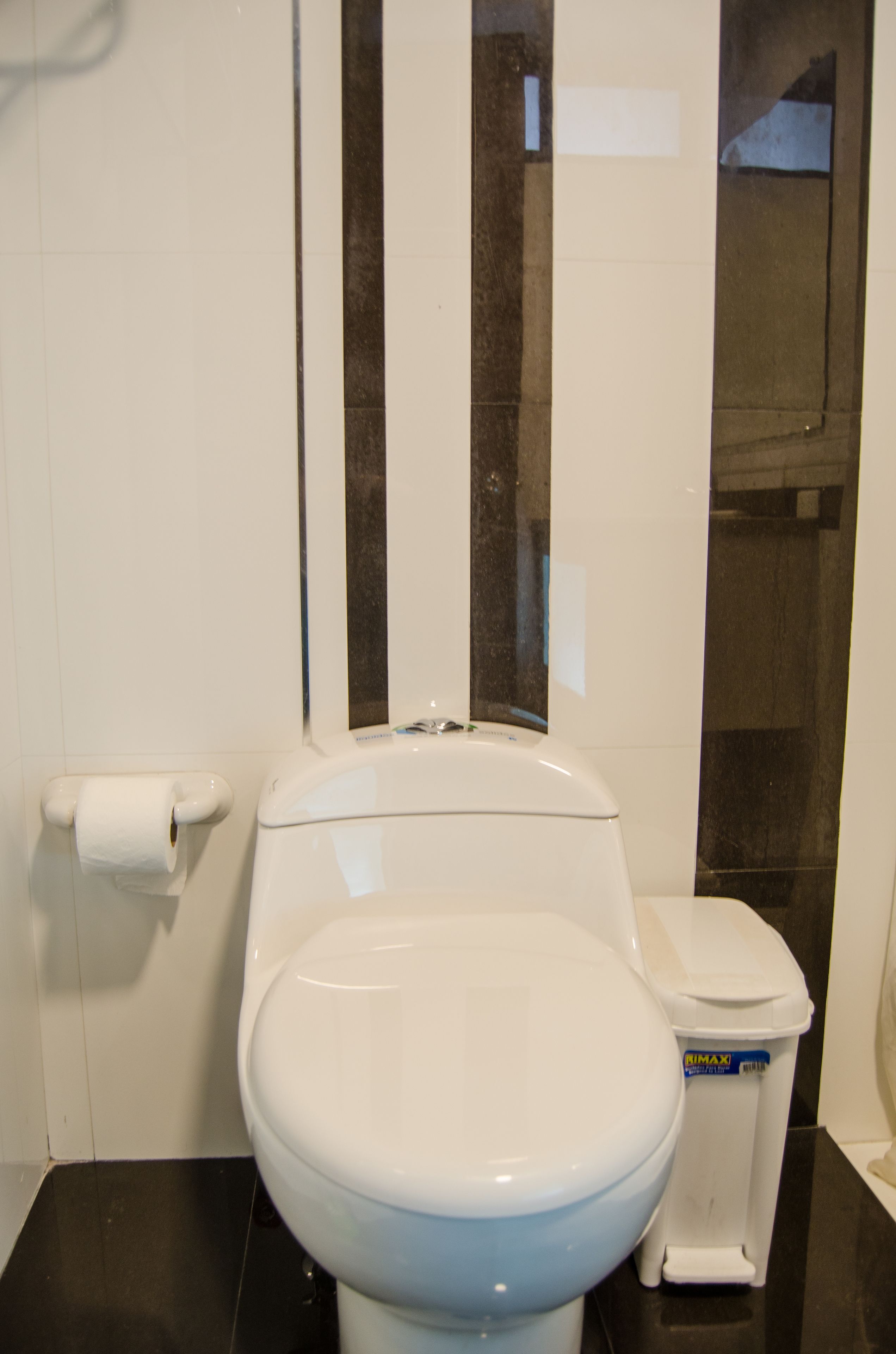 A white toilet with a white lid, a white toilet paper holder, and a white trash bin with a blue label.