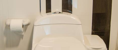 A white toilet with a white lid, a white toilet paper holder, and a white trash bin with a blue label.