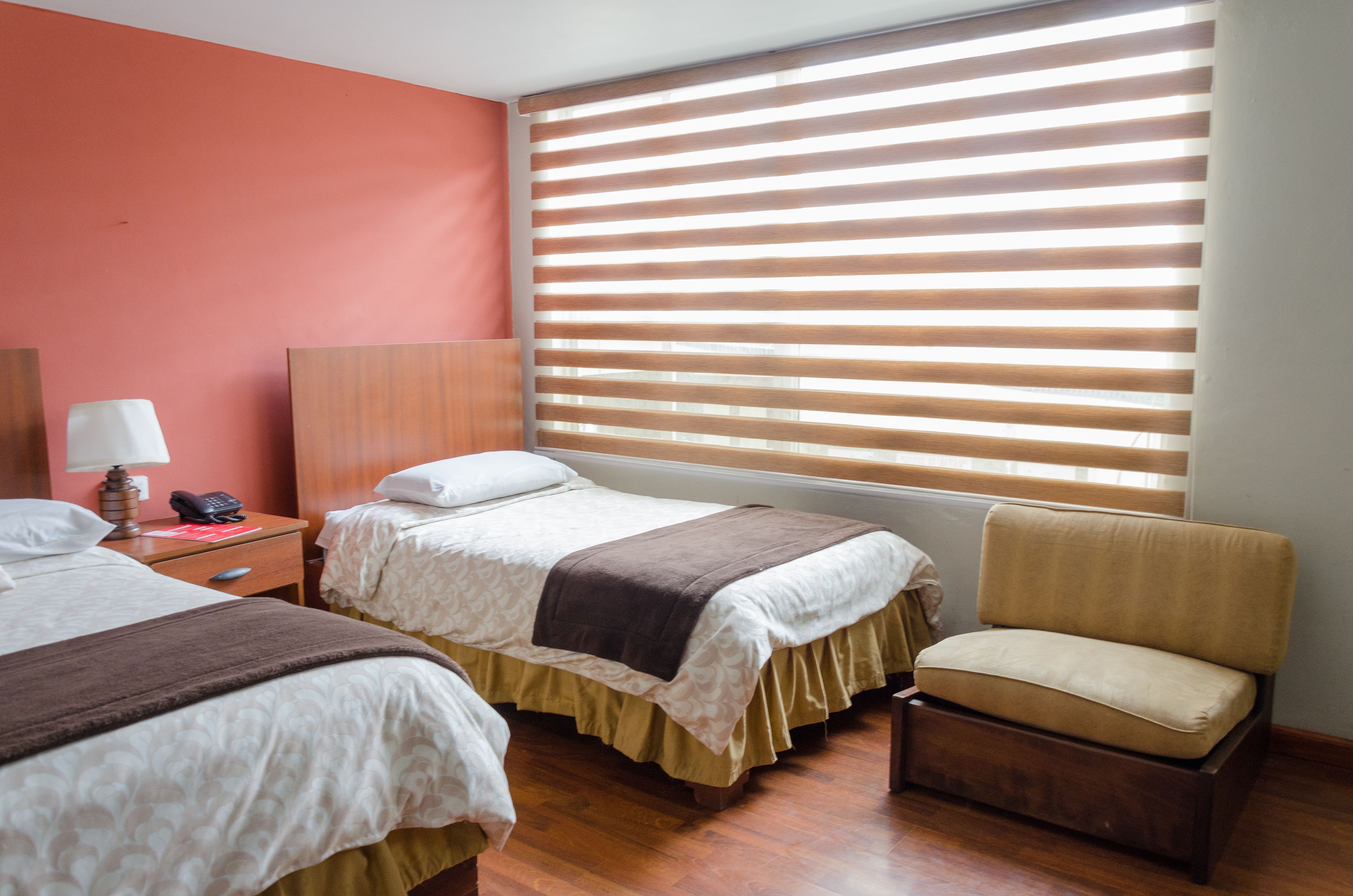 Une chambre d’hôtel avec deux lits, une chaise, une table de chevet avec une lampe et une fenêtre avec des stores.