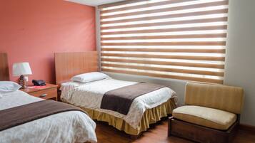 Una habitación de hotel con dos camas, una silla, una mesita de noche con lámpara y una ventana con persianas.