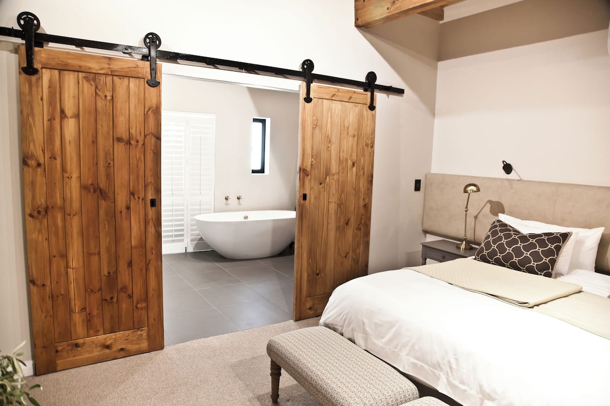 A bedroom with a large bed, a bench, a wooden sliding door, and a view into the bathroom.