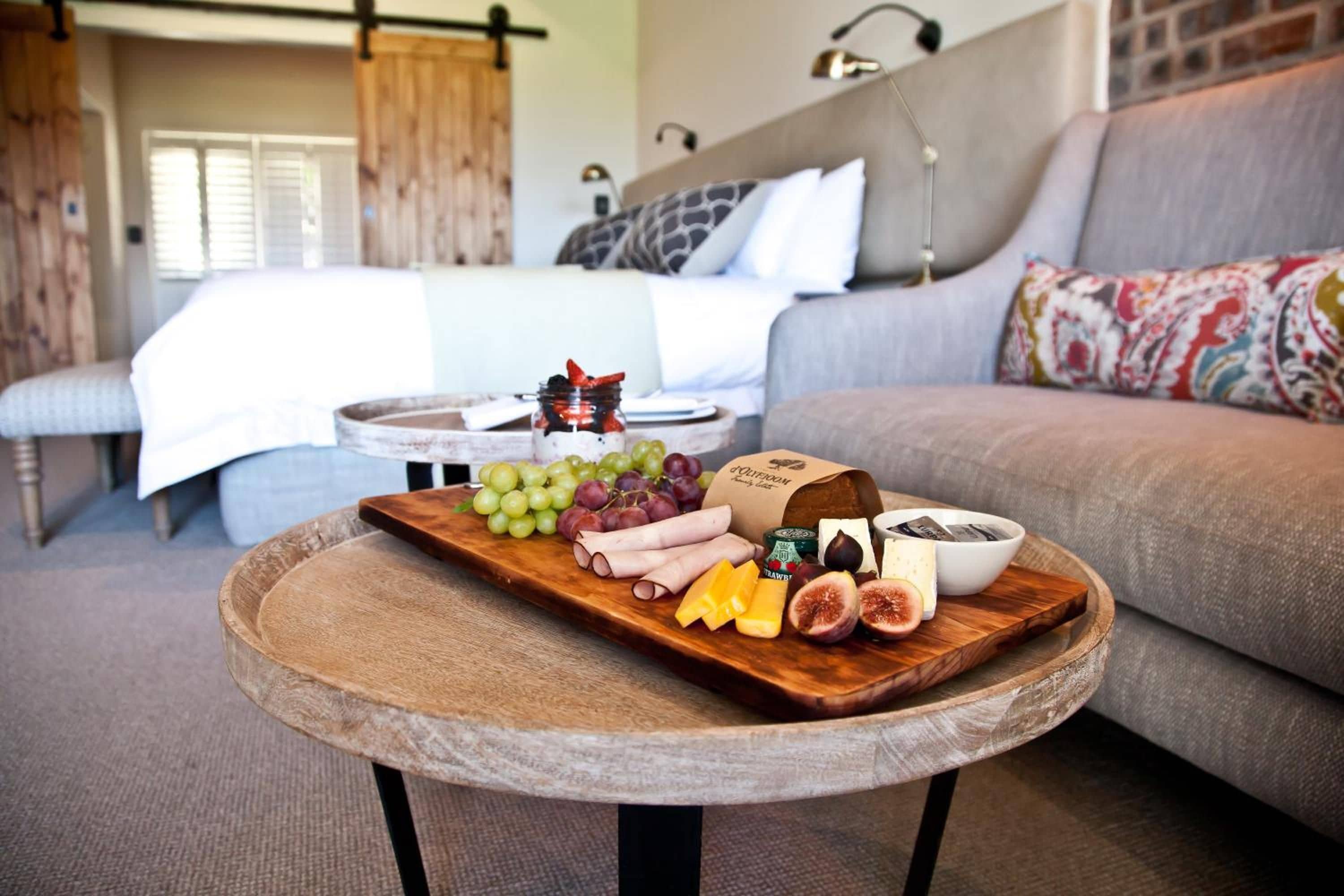 A room with a bed, a sofa, and a round table with a wooden board displaying cheese, grapes, and figs.