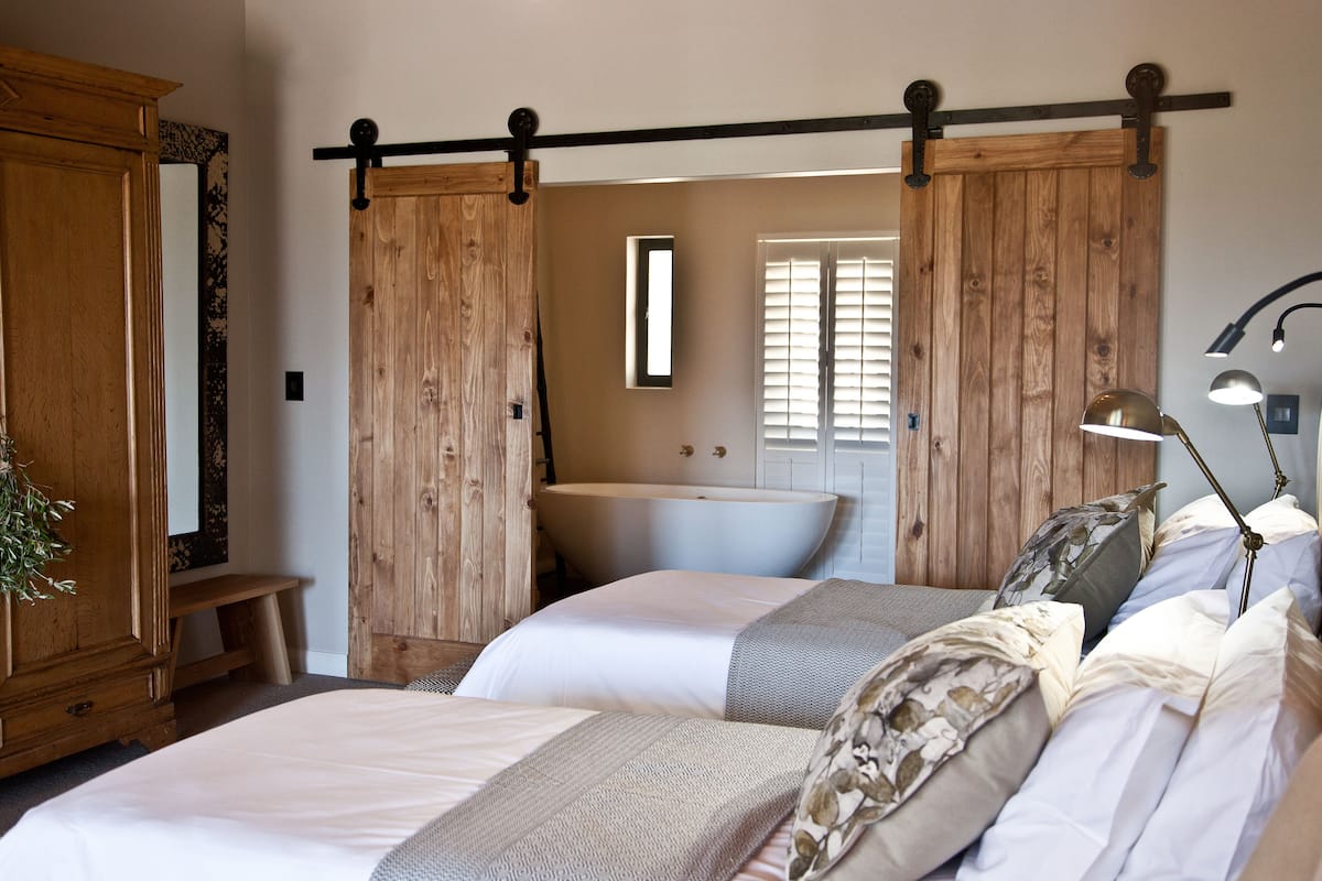 A bedroom with two beds, a wooden wardrobe, a large mirror, a sink, and a hanging light fixture.