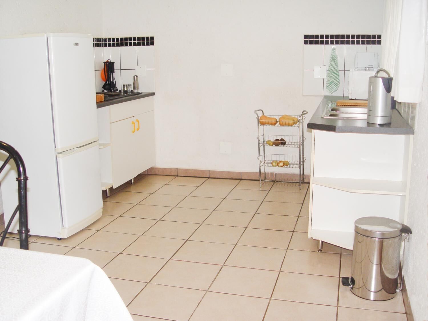 Una cocina pequeña y limpia, con armarios blancos, un refrigerador blanco, un lavamanos y una estantería metálica con utensilios de cocina.
