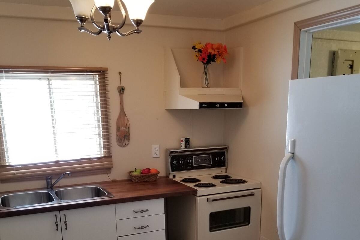A kitchen with white cabinets, a stove, a refrigerator, a window with blinds, and a chandelier.