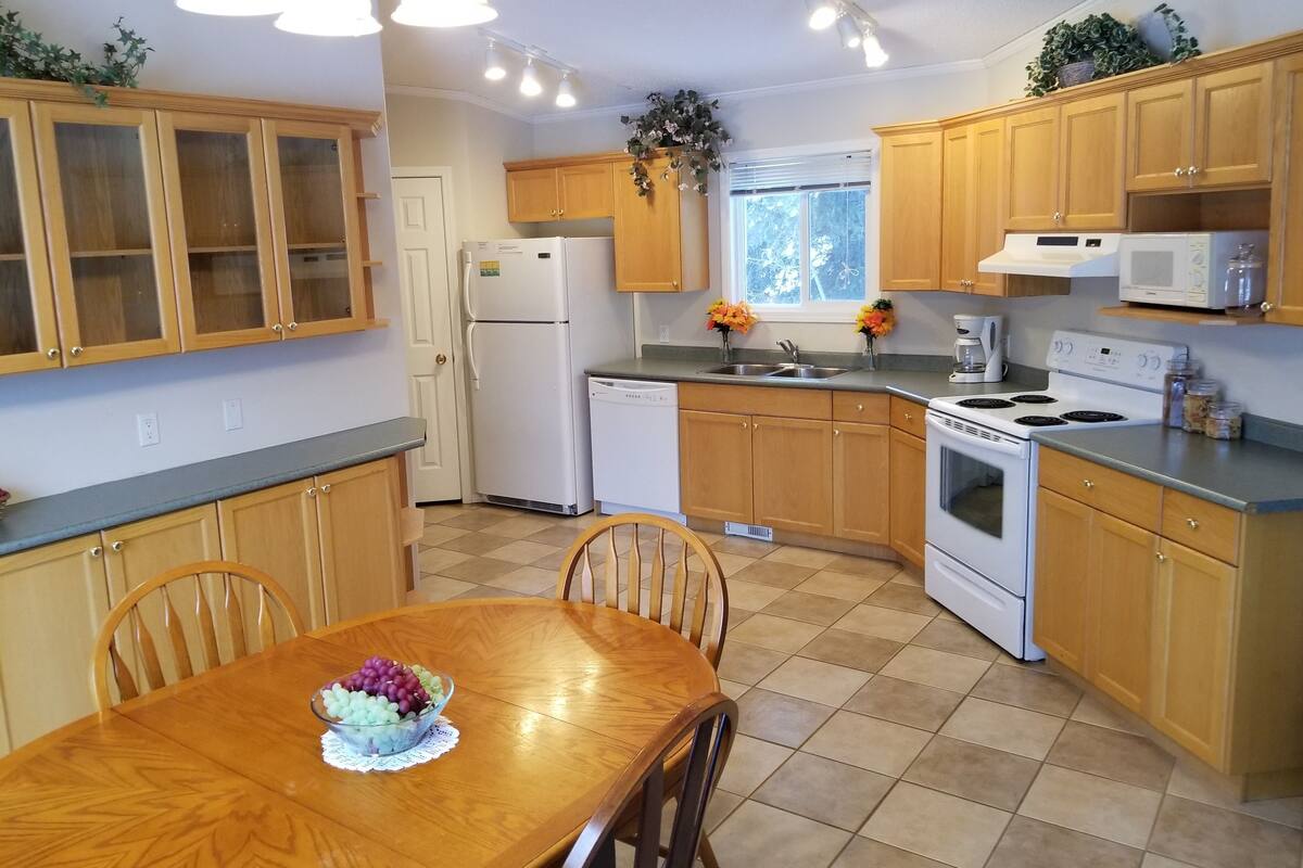 A kitchen with wooden cabinets, a refrigerator, a dishwasher, a stove, and a sink. There is a dining table with chairs and a bowl of fruit.